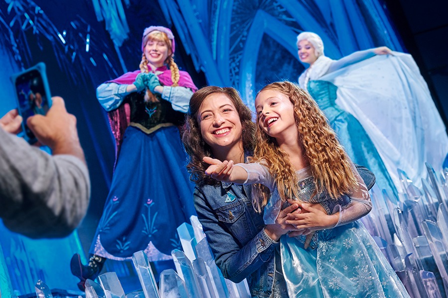 An adult and child wearing an Elsa costume smiling for a photo with Elsa and Anna from Frozen.