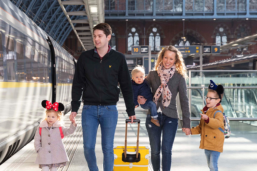 Family walking along a train platform beside a high-speed train, pulling a yellow suitcase inside a large, modern station.
