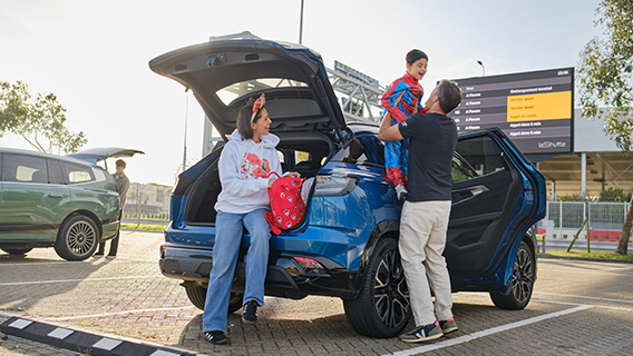 A similing woman wearing Disney merchandise sits on an open car boot as a man lifts a child in a Spider-Man costume in a car park.